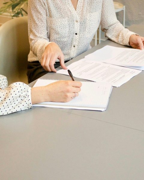woman signing on white printer paper beside woman about to touch the documents