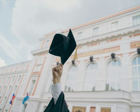 A hand holding a black graduation cap aloft before a large university building