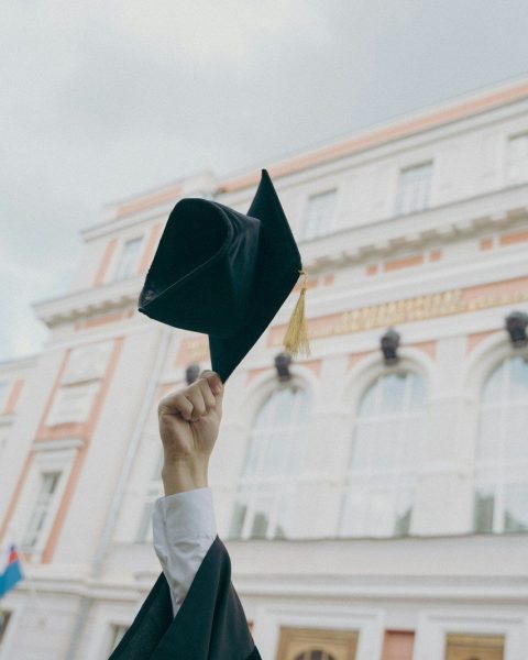A hand holding a black graduation cap aloft before a large university building
